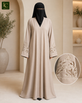 A woman modeling a long beige abaya with embroidered details on the sleeves, paired with a black face veil, in a warm neutral interior setting. An inset circle shows a close-up of the sleeve embroidery.