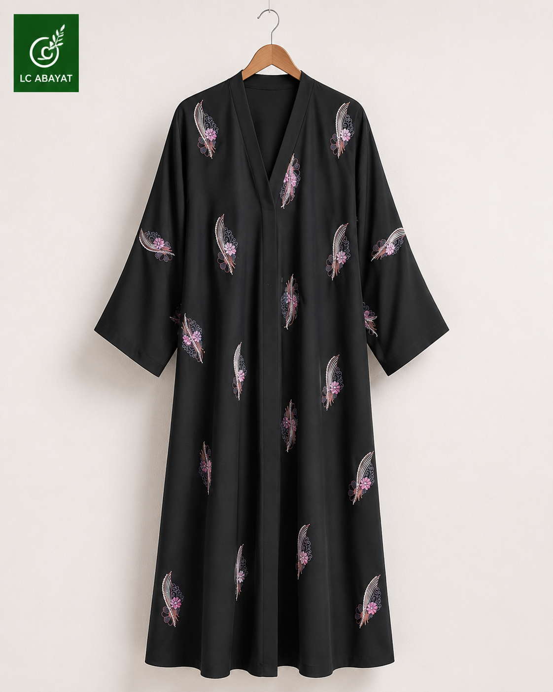 A black Abaya with wide full sleeves and a V-neckline, decorated with scattered pink and gray feather patterns, hanging on a wooden hanger against a light beige wall. The LC ABAYAT brand logo is visible in the top-left corner.