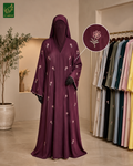 A woman wearing a long, floor-length deep purple abaya with white floral embroidery and a matching hood, standing in a boutique. An inset circle highlights the embroidery detail on the fabric.
