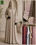 A woman modeling a long beige abaya with a matching head covering in a softly lit indoor setting. The abaya has long sleeves with pink floral embroidery near the cuffs.