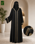 A woman modeling a black abaya with gold embroidered trim, shown in a warm indoor setting with an archway and potted plant. A circular inset highlights the gold embroidery detail on the sleeve.
