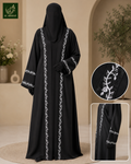 A woman modeling a black abaya with white leafy vine embroidery along the front and sleeve cuffs, shown in a warm beige indoor setting. Two inset close-ups on the right show detailed embroidery and fabric texture.