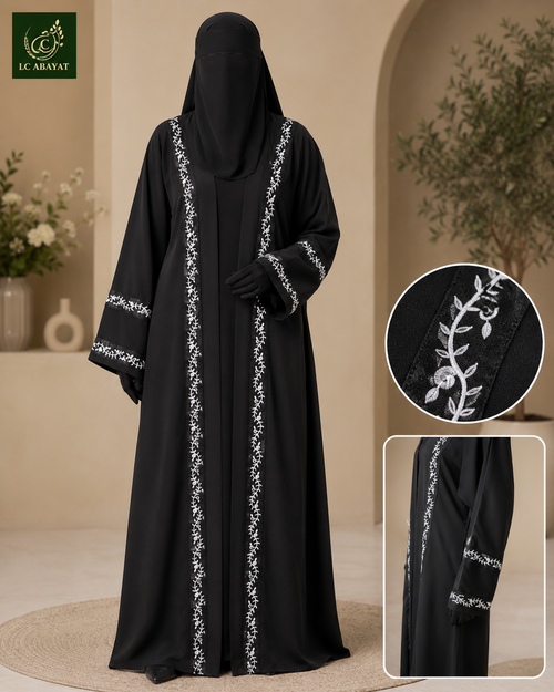A woman modeling a black abaya with white leafy vine embroidery along the front and sleeve cuffs, shown in a warm beige indoor setting. Two inset close-ups on the right show detailed embroidery and fabric texture.