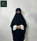 A woman wearing a black full-face Aalima Niqab and a white abaya stands against a plain light gray wall. The brand logo 'IC Abayas' is visible in the top-left corner.