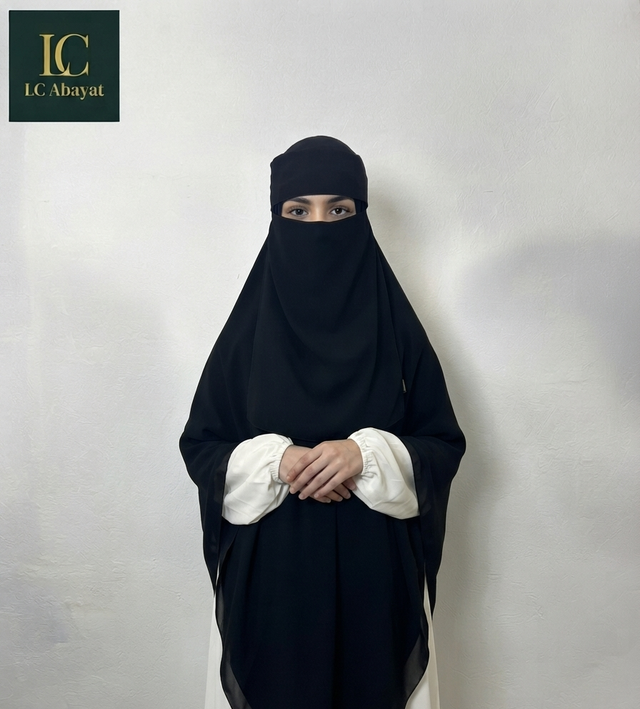 A woman wearing a black full-face Aalima Niqab and a white abaya stands against a plain light gray wall. The brand logo 'IC Abayas' is visible in the top-left corner.