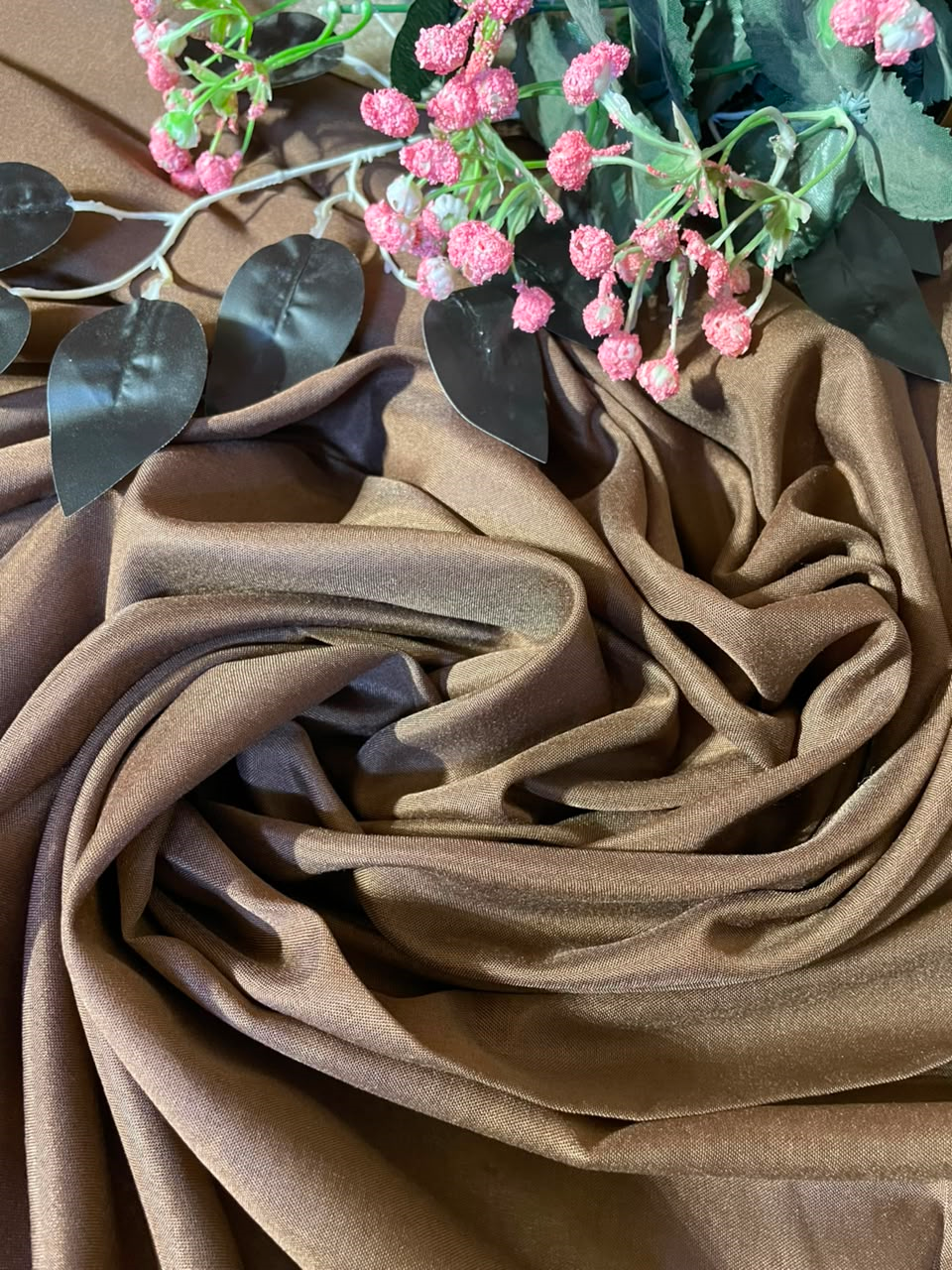 Brown draped fabric with soft folds, styled with pink flowers and dark green leaves on a matching brown background.