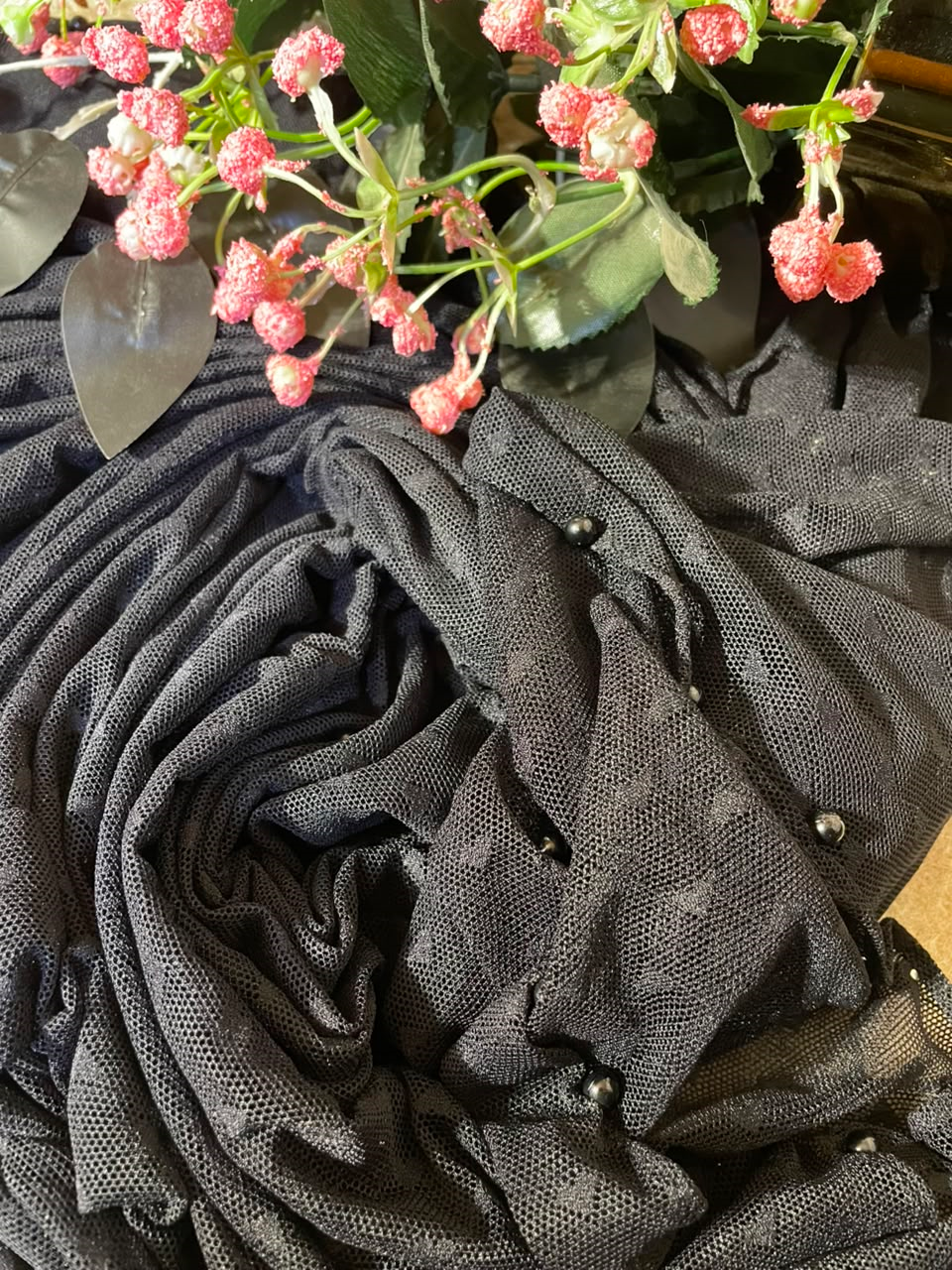 A close-up of a dark charcoal gray sheer mesh fabric draped in soft folds, decorated with small silver beads and accented by pink flowers and green leaves at the top.