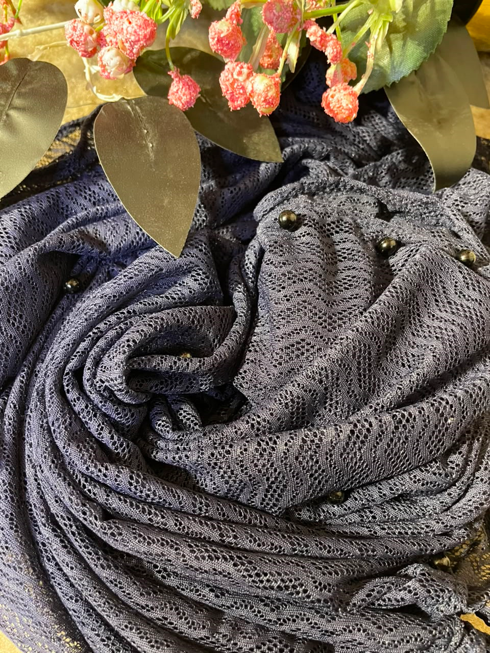 A close-up of a dark grey open-knit scarf or shawl with small dark bead accents, styled with decorative leaves and pink flowers at the top.