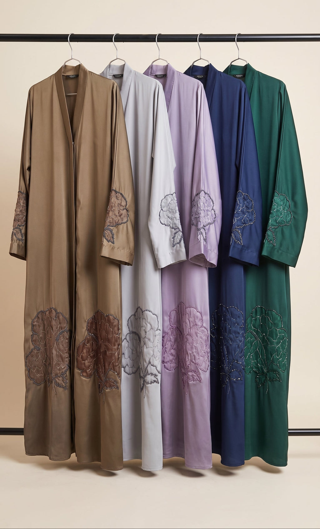 Four long open-front handmade beadwork abayas hanging on a black rack against a plain beige wall. From left to right, the colors are taupe-brown, light gray, lavender, navy blue, and deep green.