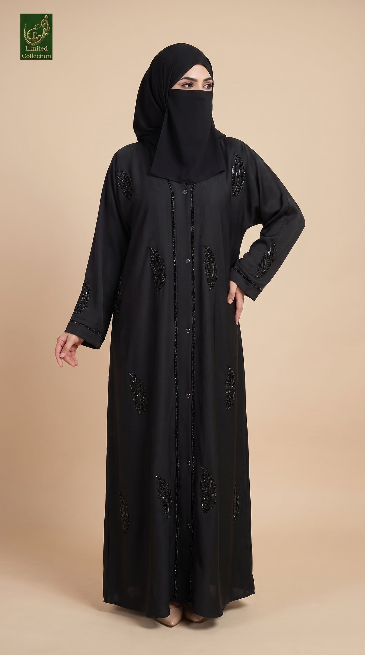 A woman modeling a long black abaya with matching black hijab, standing against a solid beige studio background. The abaya features subtle embroidered details on the front and sleeves.