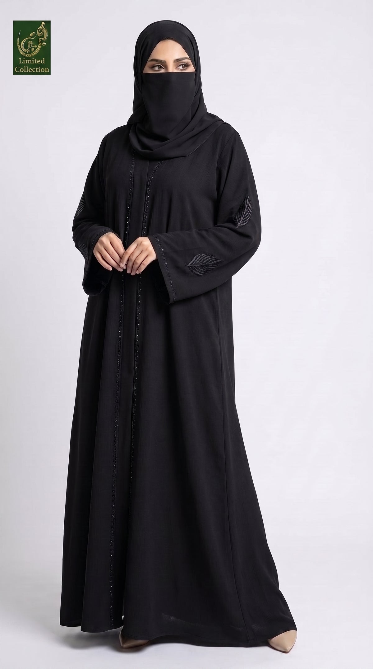 A woman wearing a long black abaya and matching black hijab, standing against a light gray studio background. A small green and gold logo appears in the top-left corner.