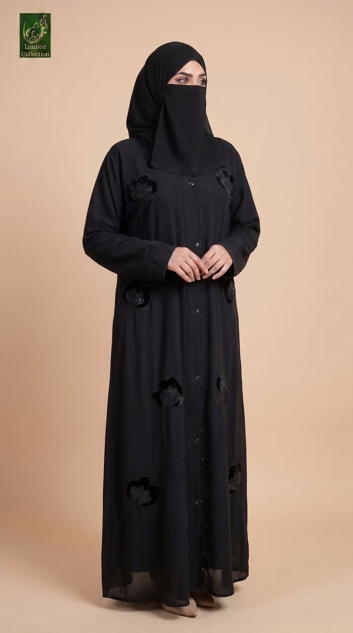 A woman wearing a black abaya with floral applique and a matching black hijab, standing against a solid beige studio background. A green logo for 'Abayat Limited Collection' is visible in the top-left corner.