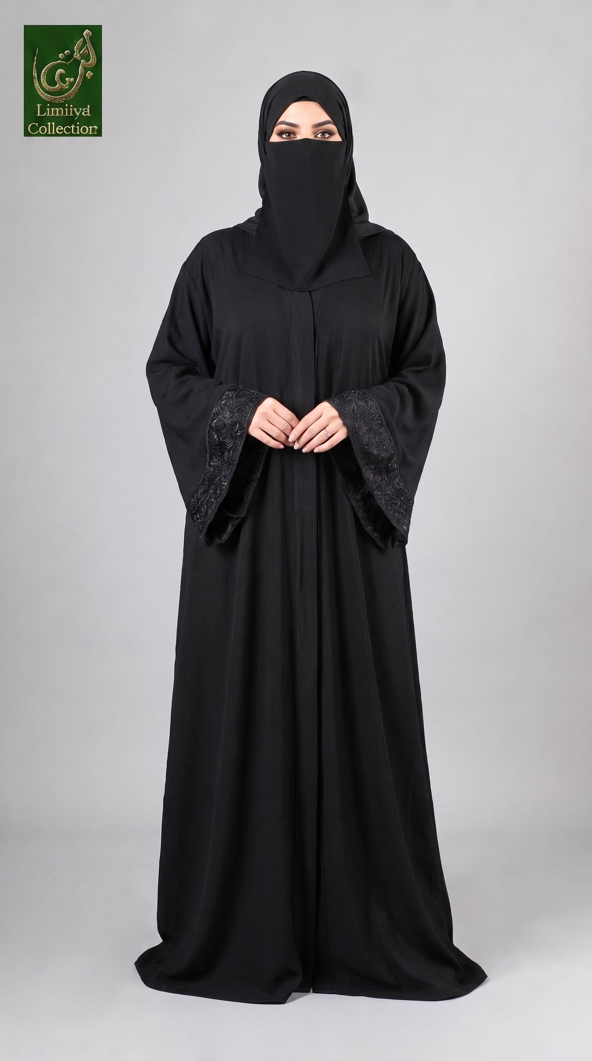 A woman wearing a full-length black abaya with wide sleeves and a matching black face covering, standing against a light gray studio background. A green and gold 'Abayat Limited Collection' logo is visible in the top-left corner.