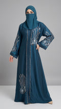 A woman modeling a long teal abaya dress with white floral embroidery and a matching teal face covering, standing against a neutral gray studio background.