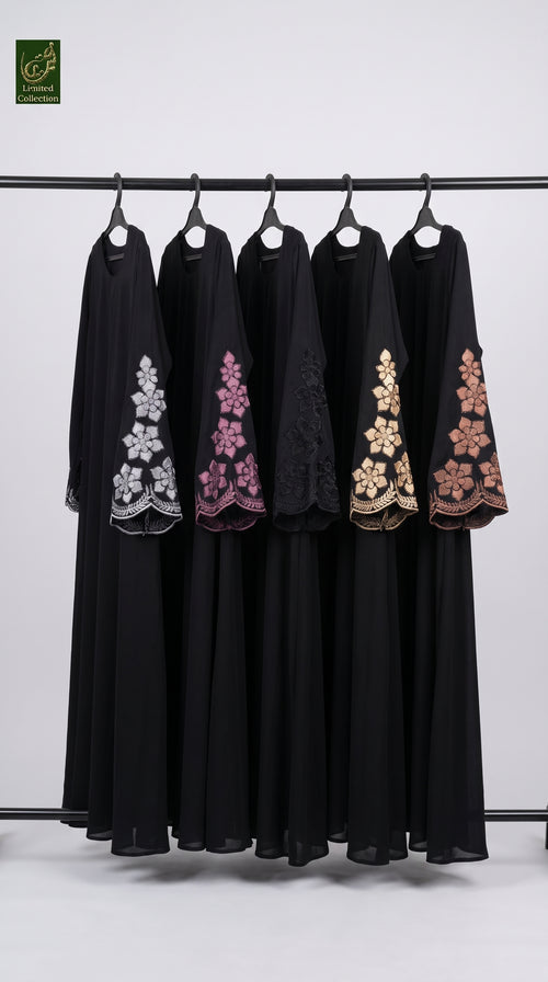 Five black abayas hanging on a black clothing rack against a light gray background. Each abaya features a different colored floral embroidery design on the sleeve: silver, pink, black, gold, and brown.