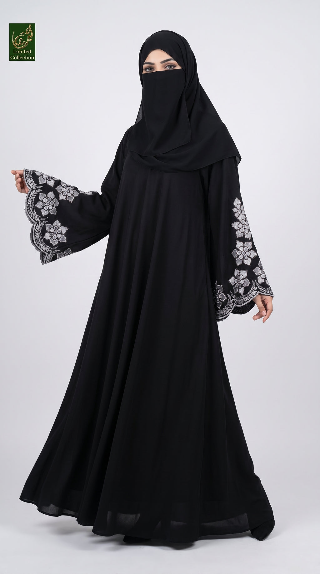 A woman modeling a black abaya with long wide sleeves featuring grey floral embroidery, paired with a matching black hijab. She is standing against a light gray studio background.