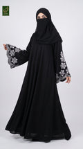 A woman modeling a black abaya with long wide sleeves featuring grey floral embroidery, paired with a matching black hijab. She is standing against a light gray studio background.