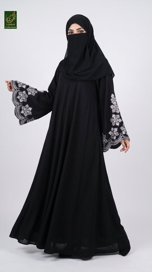 A woman modeling a black abaya with long wide sleeves featuring grey floral embroidery, paired with a matching black hijab. She is standing against a light gray studio background.