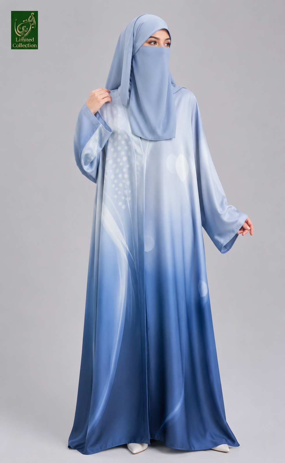 A woman wearing a flowing abaya and matching hijab in shades of blue with a gradient effect, standing against a solid gray studio background. A small green logo is visible in the top-left corner.