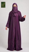 A woman wearing a long, flowing purple abaya with matching hijab and face veil, standing against a light gray studio background. The abaya has subtle embroidered detailing near the lower hem.