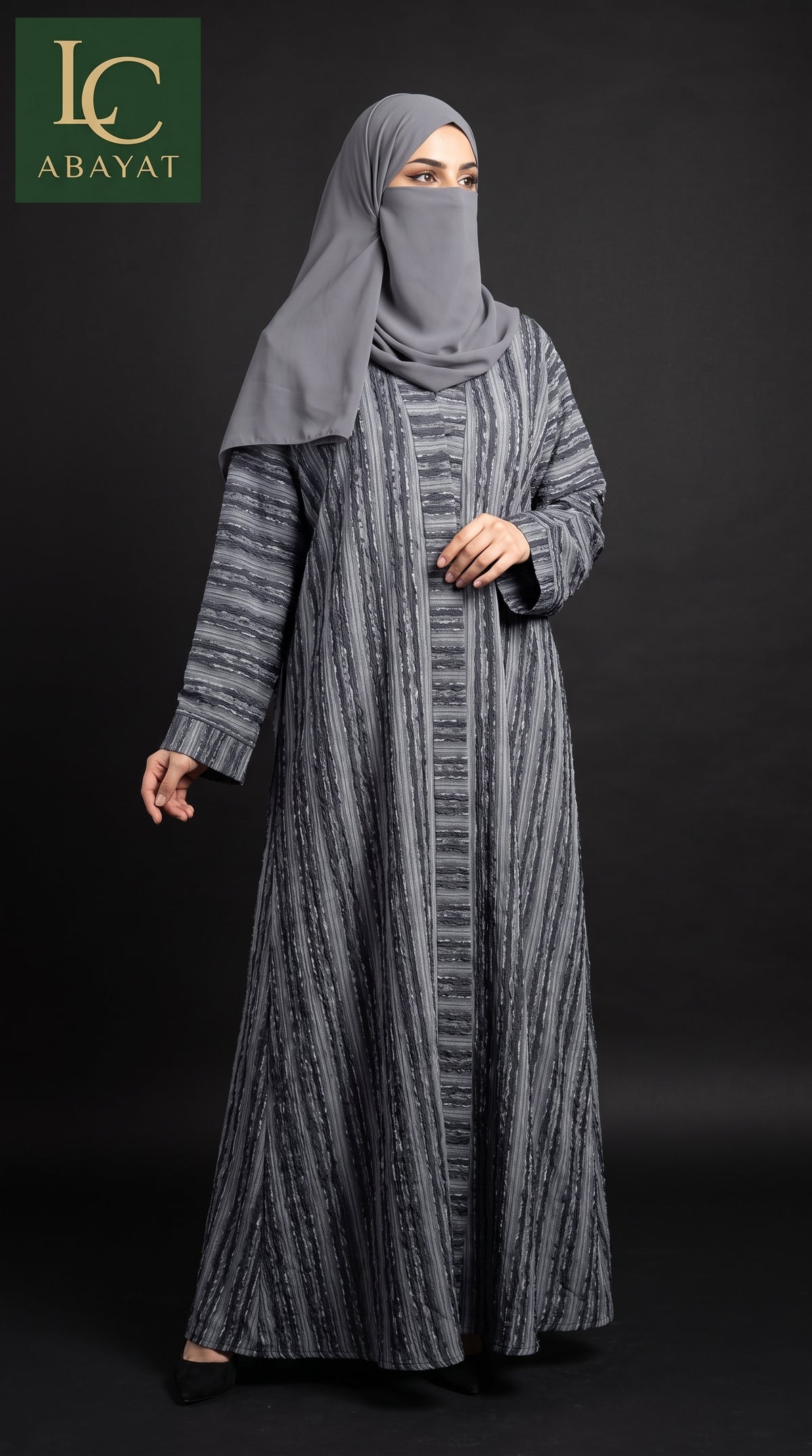A woman modeling a long gray abaya dress with a vertical textured stripe pattern, paired with a matching gray hijab and face veil, set against a dark studio background. The 'LC ABAYAT' brand logo is visible in the top-left corner.
