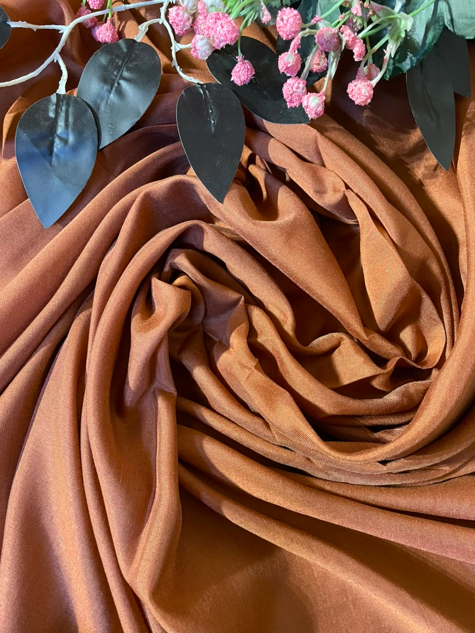 A draped piece of warm brown fabric with soft folds, styled with artificial dark green leaves and pink flowers at the top edge.