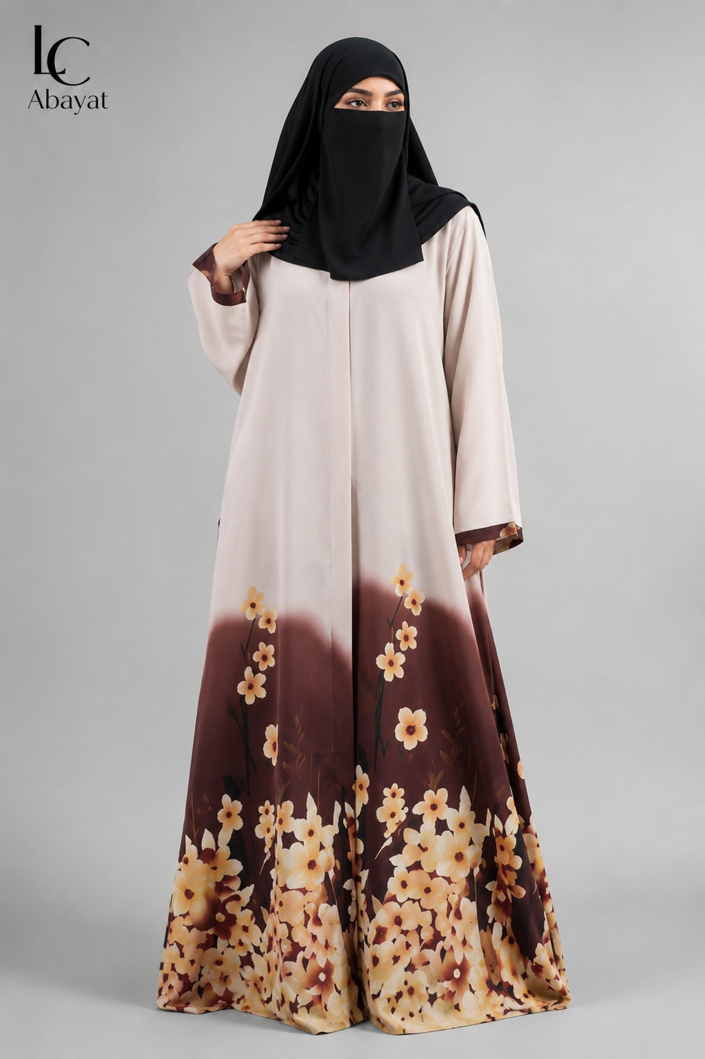 A woman wearing a long modest front open floral print abaya dress with a black hijab, shown against a plain light gray studio background. The abaya is light beige at the top and fades into a dark brown floral print at the bottom, with cream and tan flowers throughout.