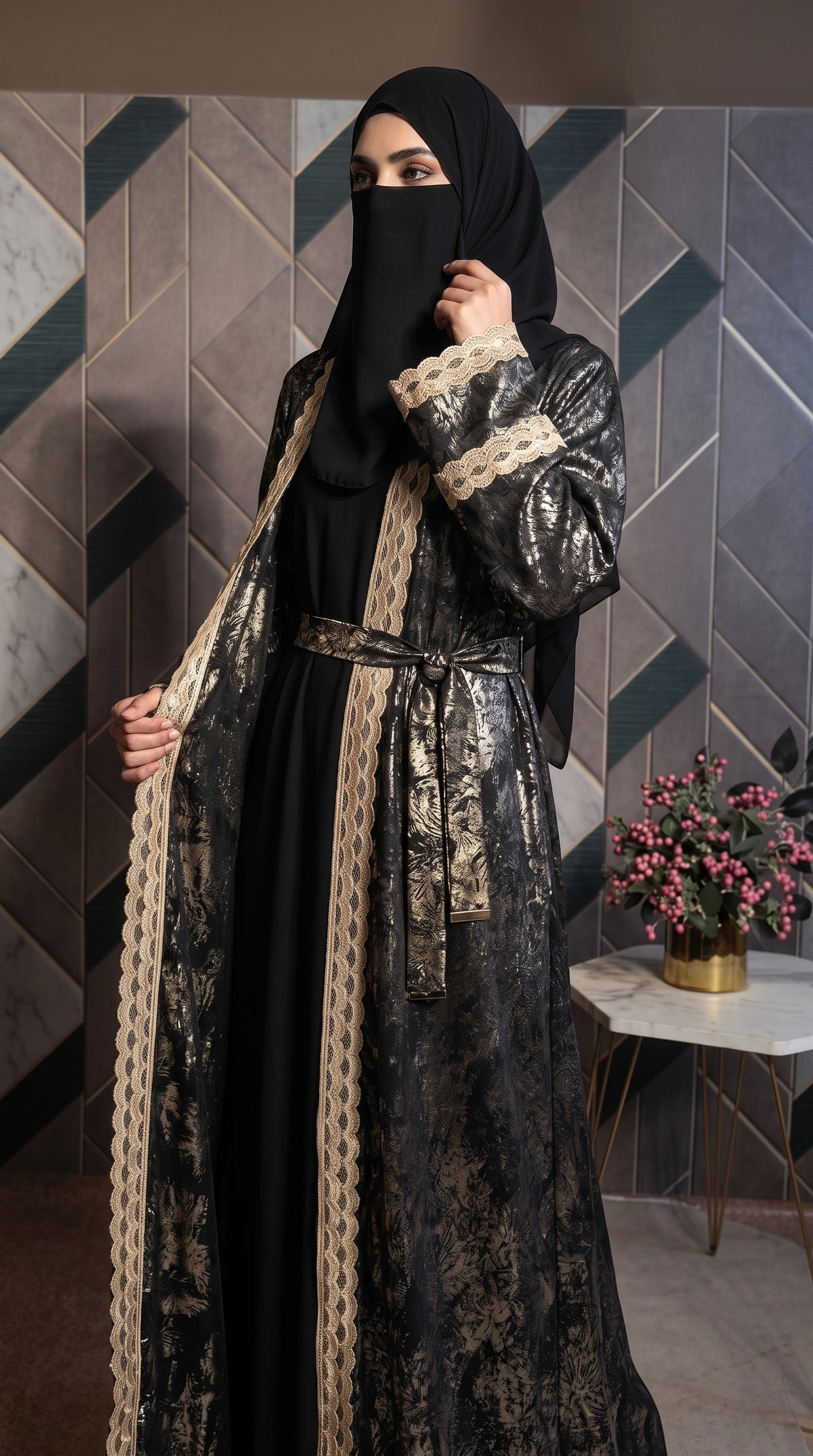 A woman wearing a black abaya with a black hijab, featuring a black and gold patterned outer layer with beige lace trim, standing in front of a geometric paneled wall.