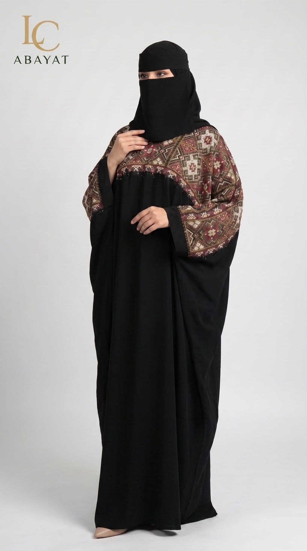 A woman modeling a black abaya with ornate brown and beige patterned sleeves, paired with a black face veil, against a light gray studio background. The LC Abayat logo is visible in the top left corner.