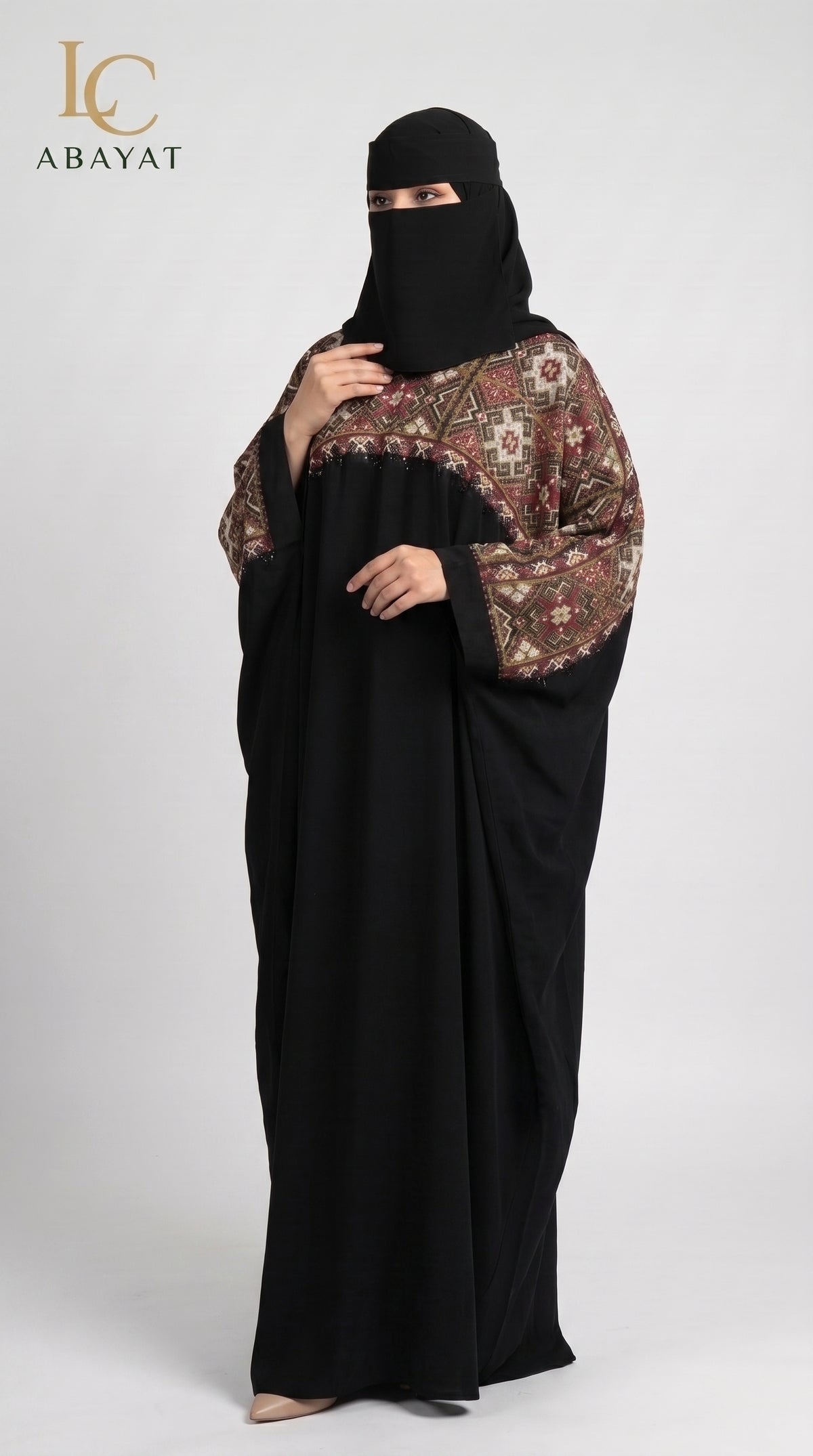 A woman modeling a black abaya with ornate brown and beige patterned sleeves, paired with a black face veil, against a light gray studio background. The LC Abayat logo is visible in the top left corner.