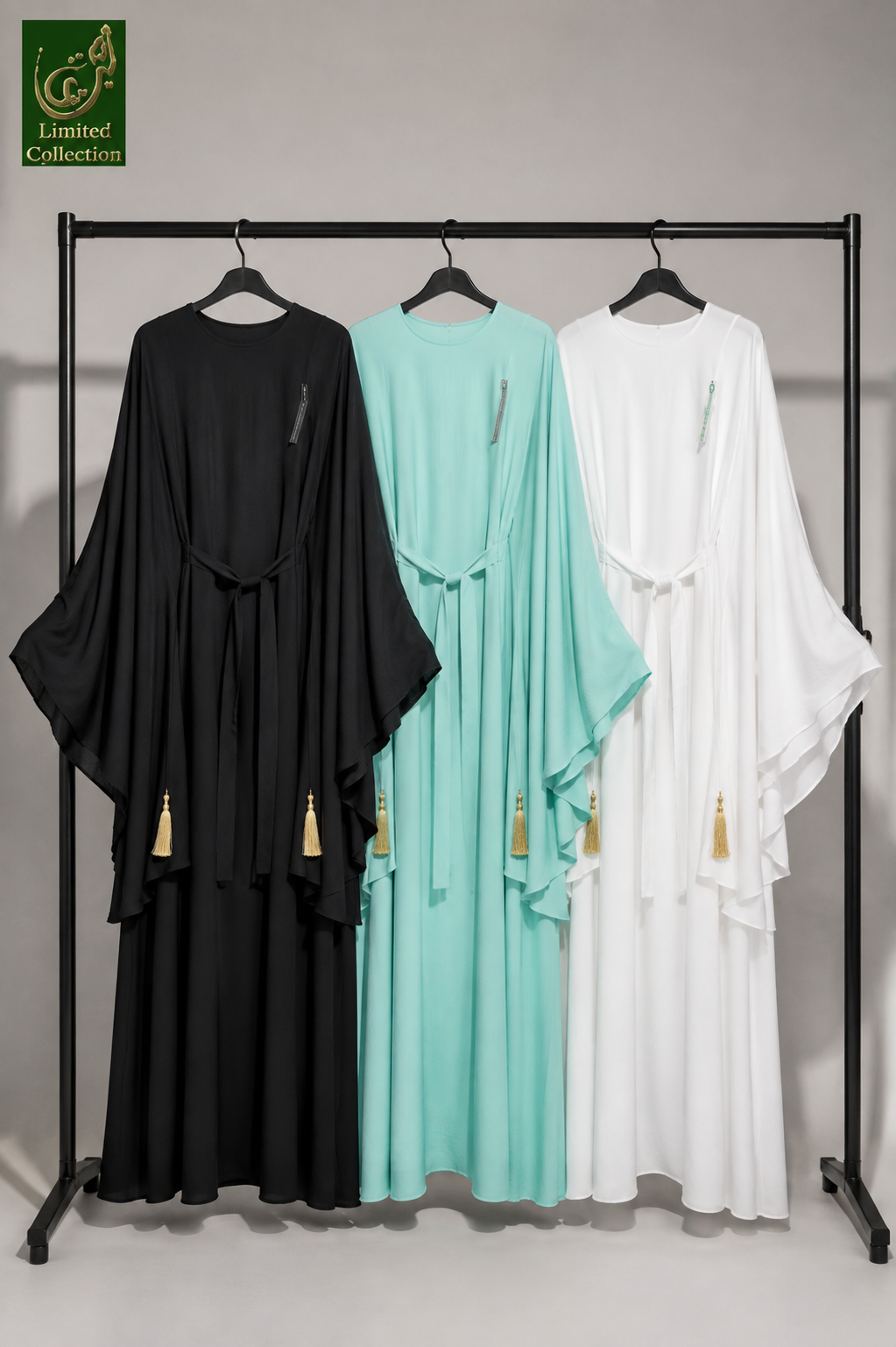 Three long flowing maxi abayas in black, mint green, and white hang on a black clothing rack against a light gray studio background. Each abaya has wide draped sleeves, a wrap-style front, and small gold tassel accents near the lower front.