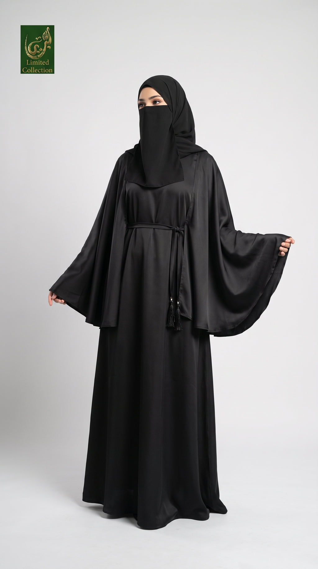 A woman modeling a black abaya with a matching black hijab and face veil, standing against a light gray studio background. The abaya has a loose, flowing silhouette with wide sleeves and a drawstring waist.