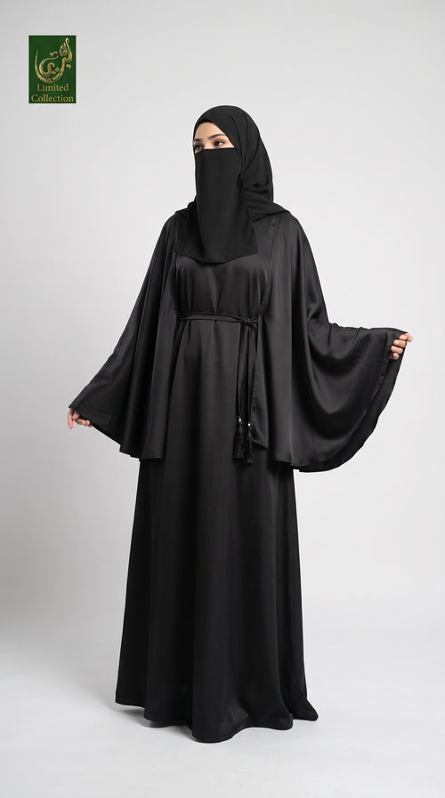 A woman modeling a black abaya with a matching black hijab and face veil, standing against a light gray studio background. The abaya has a loose, flowing silhouette with wide sleeves and a drawstring waist.