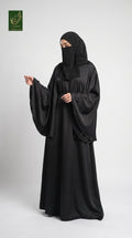 A woman modeling a black abaya and matching black hijab against a light gray studio background. A small green and gold logo appears in the top-left corner.