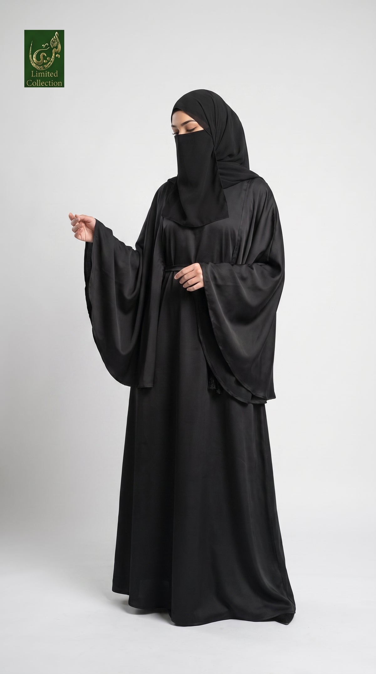 A woman modeling a black abaya and matching black hijab against a light gray studio background. A small green and gold logo appears in the top-left corner.