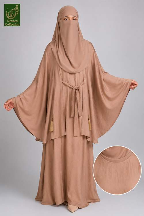 A woman modeling a modest beige abaya and matching head covering against a light gray studio background. The abaya has wide draped sleeves, a waist tie, and small tassel accents, with a circular inset showing a close-up of the fabric texture.