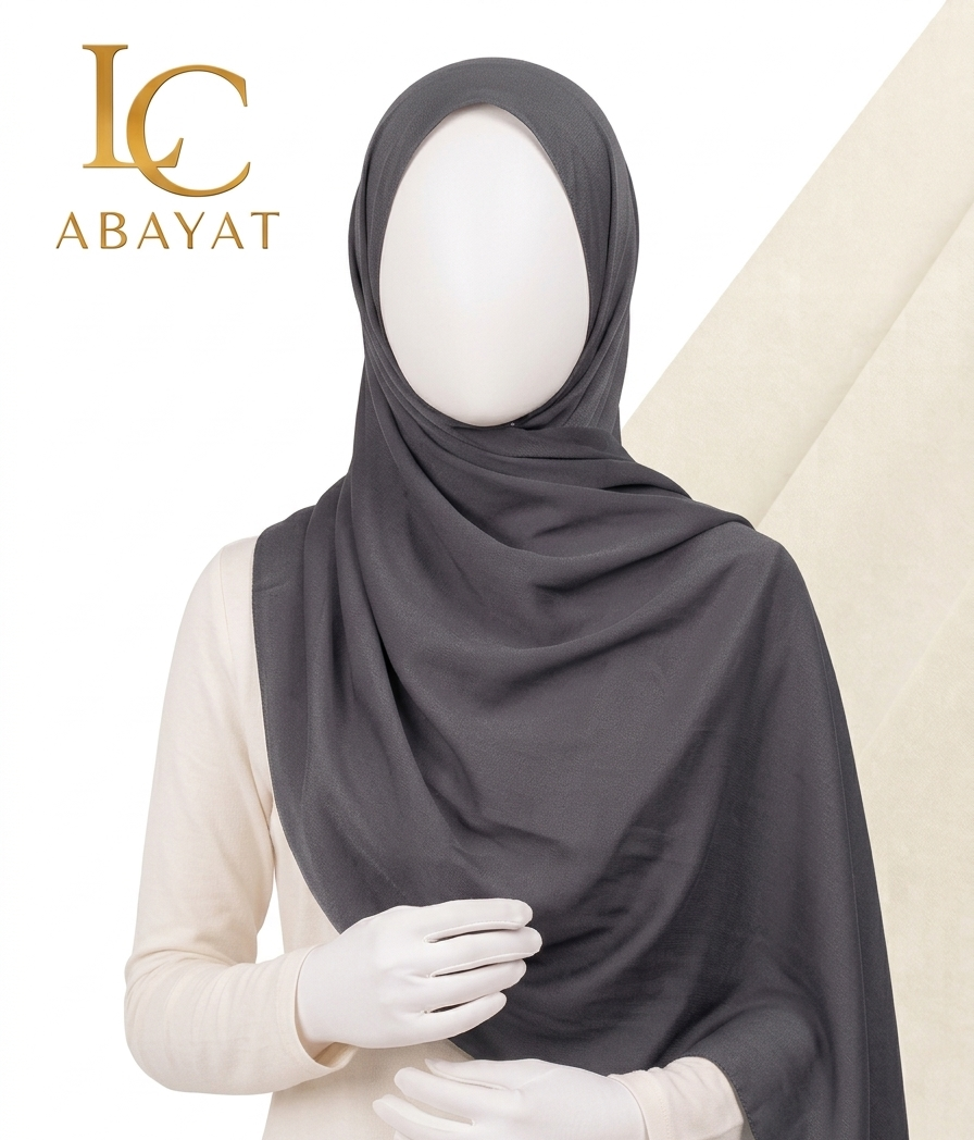 A dark charcoal gray hijab draped elegantly on a white mannequin wearing a cream-colored dress, with the 'IC ABAYAT' brand logo in gold in the top-left corner.