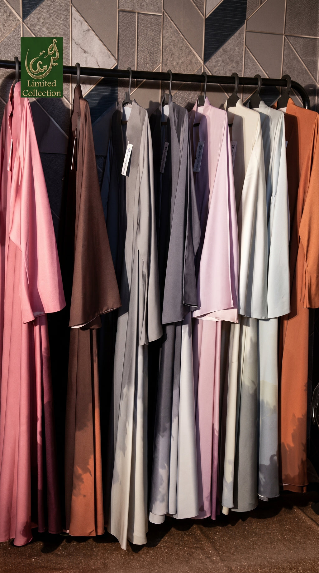 A collection of long, flowing Front Open Tie-Dye Long Flair Abayas in various colors hanging on a black rack against a geometric tiled wall. The garments include shades of pink, brown, navy, gray, lavender, light blue, and orange.