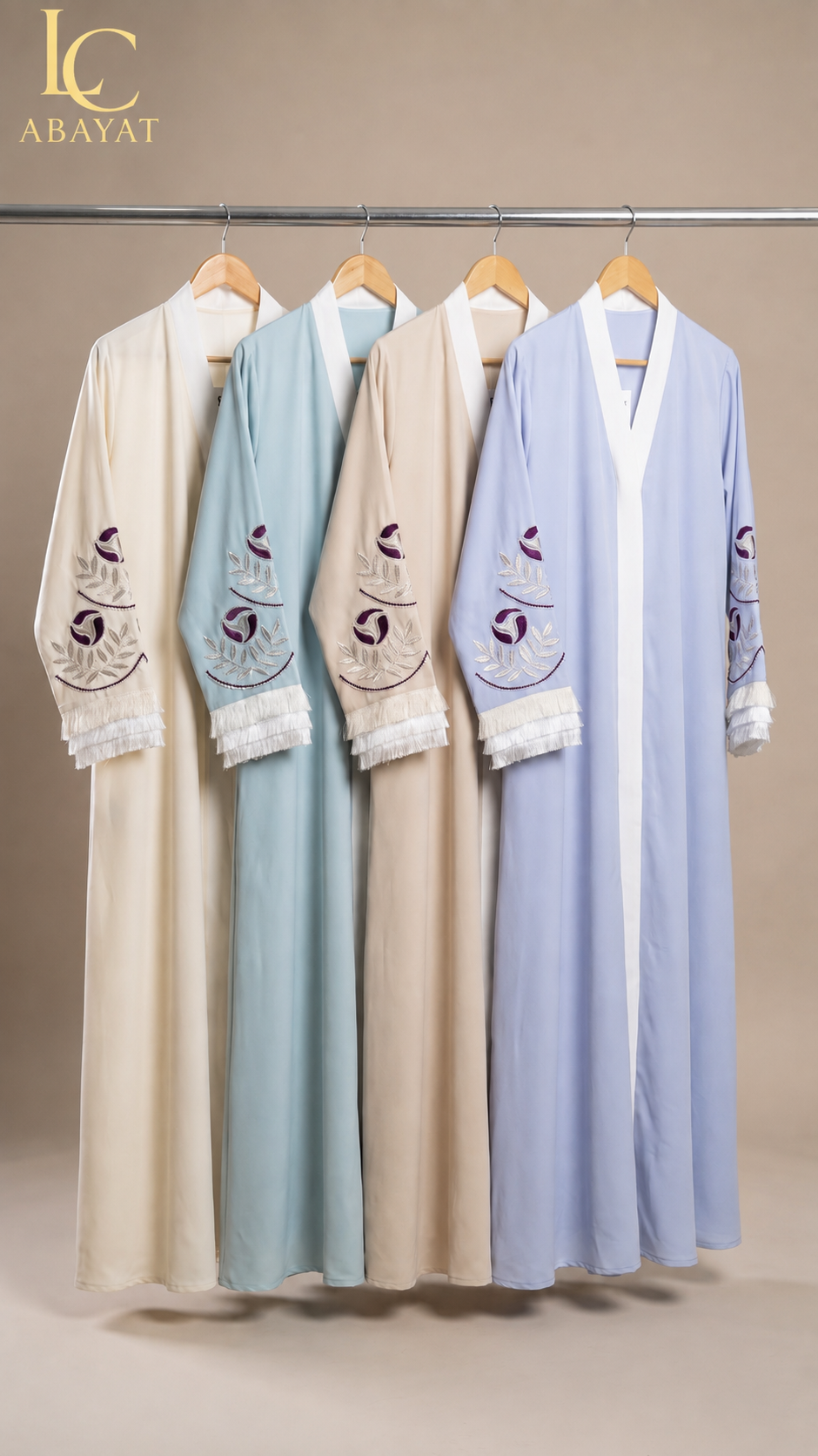 Four long open-front abaya with embroidered sleeves & fringed cuffs hanging on a metal rack against a warm beige studio background. The abayas come in cream, pale blue, beige, and light lavender-blue, each with embroidered sleeve details and white lace trim at the cuffs.