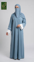 A woman modeling a light blue abaya and matching hijab set with white lace trim on the sleeves, photographed against a plain light gray studio background. A green 'Limited Collection' logo appears in the top-left corner.
