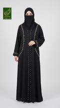 A woman wearing a long black abaya with gold decorative trim and a matching black face covering, standing against a light gray studio background. A green 'Gourmet Collection' logo appears in the top-left corner.