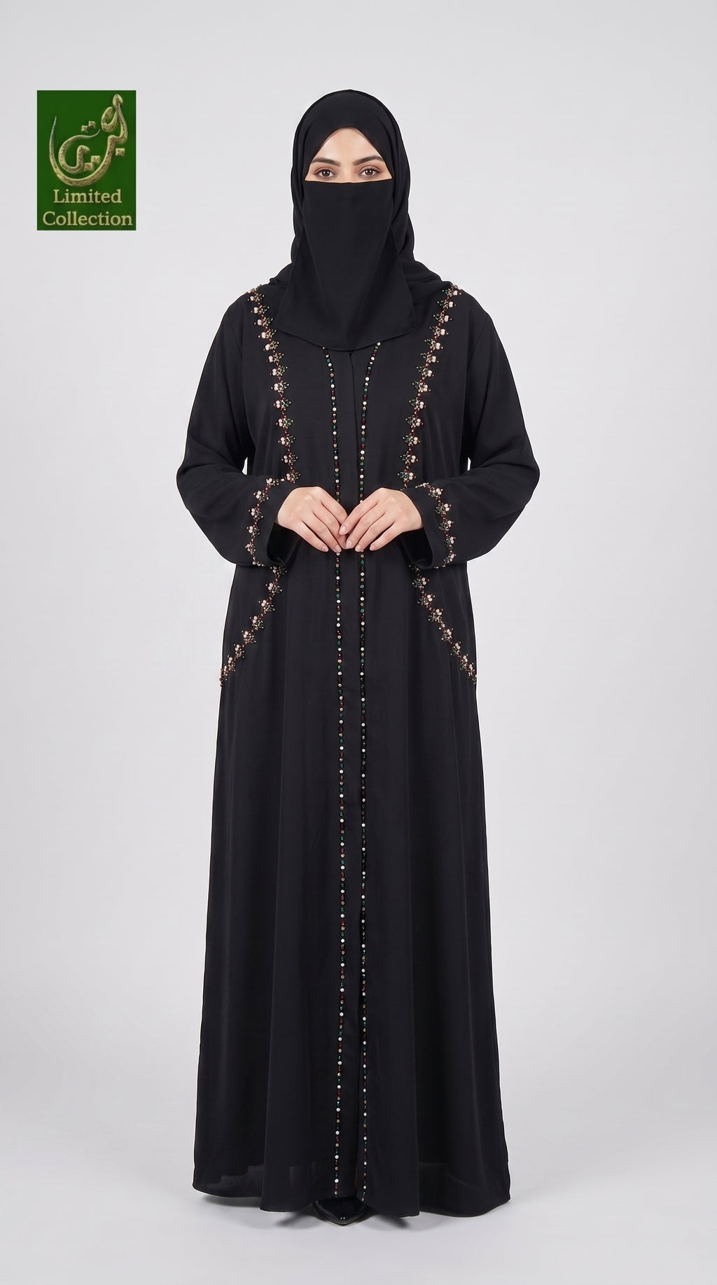 A woman wearing a long black abaya with gold button accents and a matching black hijab, standing against a light gray studio background. A green 'Abayat Limited Collection' logo is visible in the top-left corner.