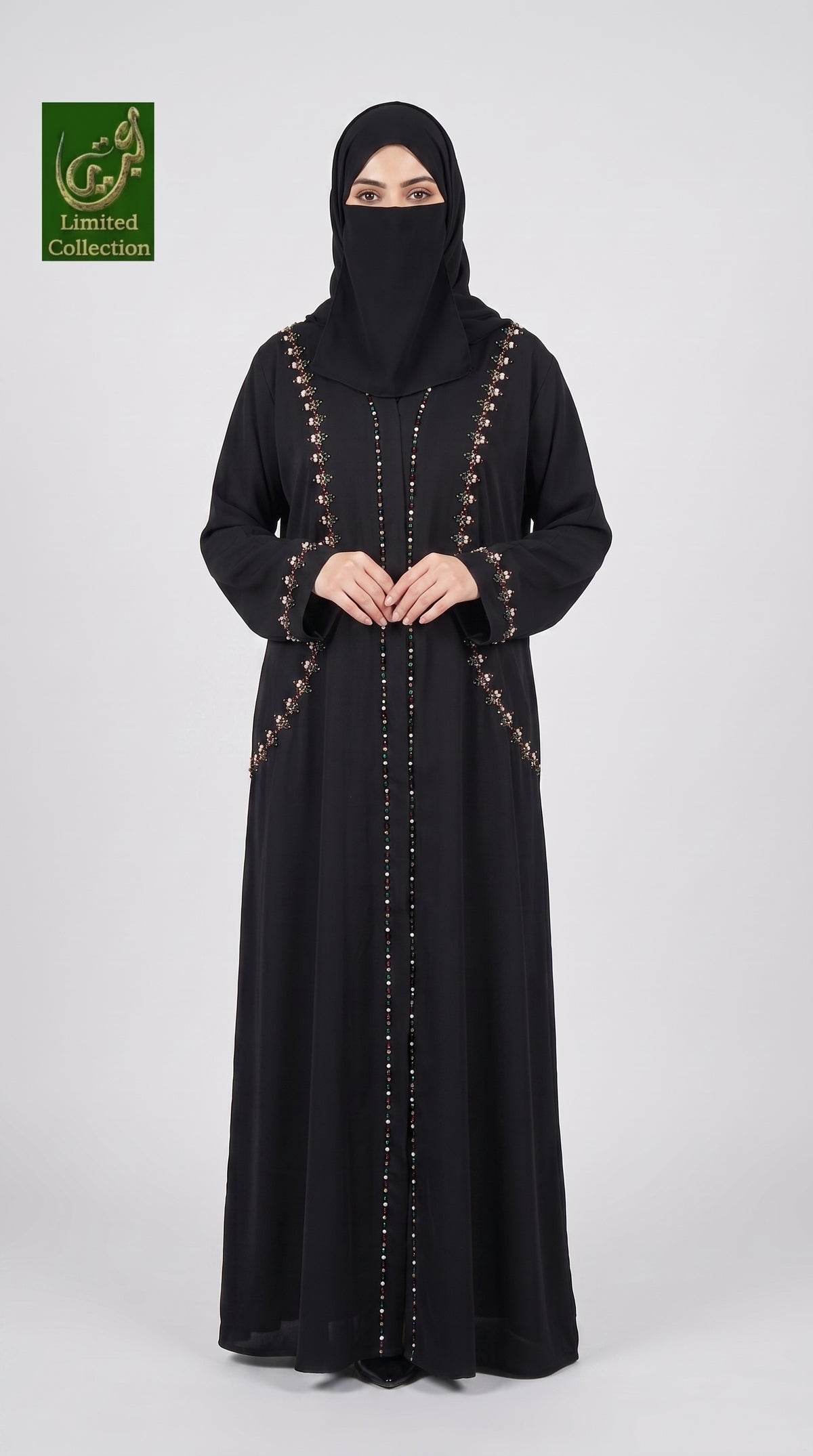 A woman wearing a long black abaya with gold button accents and a matching black hijab, standing against a light gray studio background. A green 'Abayat Limited Collection' logo is visible in the top-left corner.