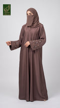 A woman modeling a long brown abaya with matching hijab, featuring embroidered detailing on the sleeves, set against a plain light gray background. A green 'GCL Limited Collection' logo is visible in the top-left corner.