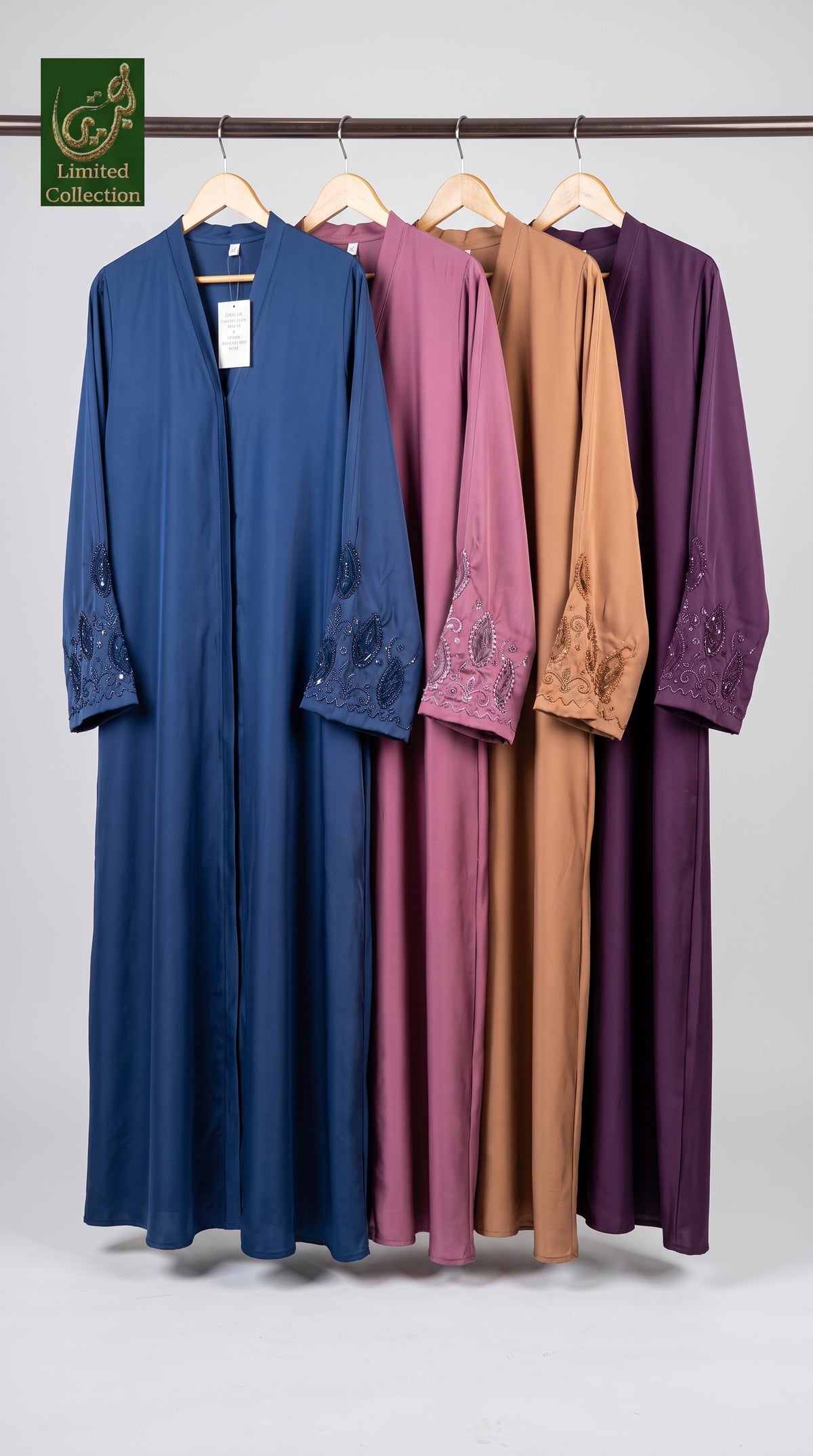 Four long modest abayas in blue, dusty pink, camel brown, and deep purple hanging on a wooden rack against a light gray background. Each abaya features embroidered detailing on the sleeves and a V-neckline.