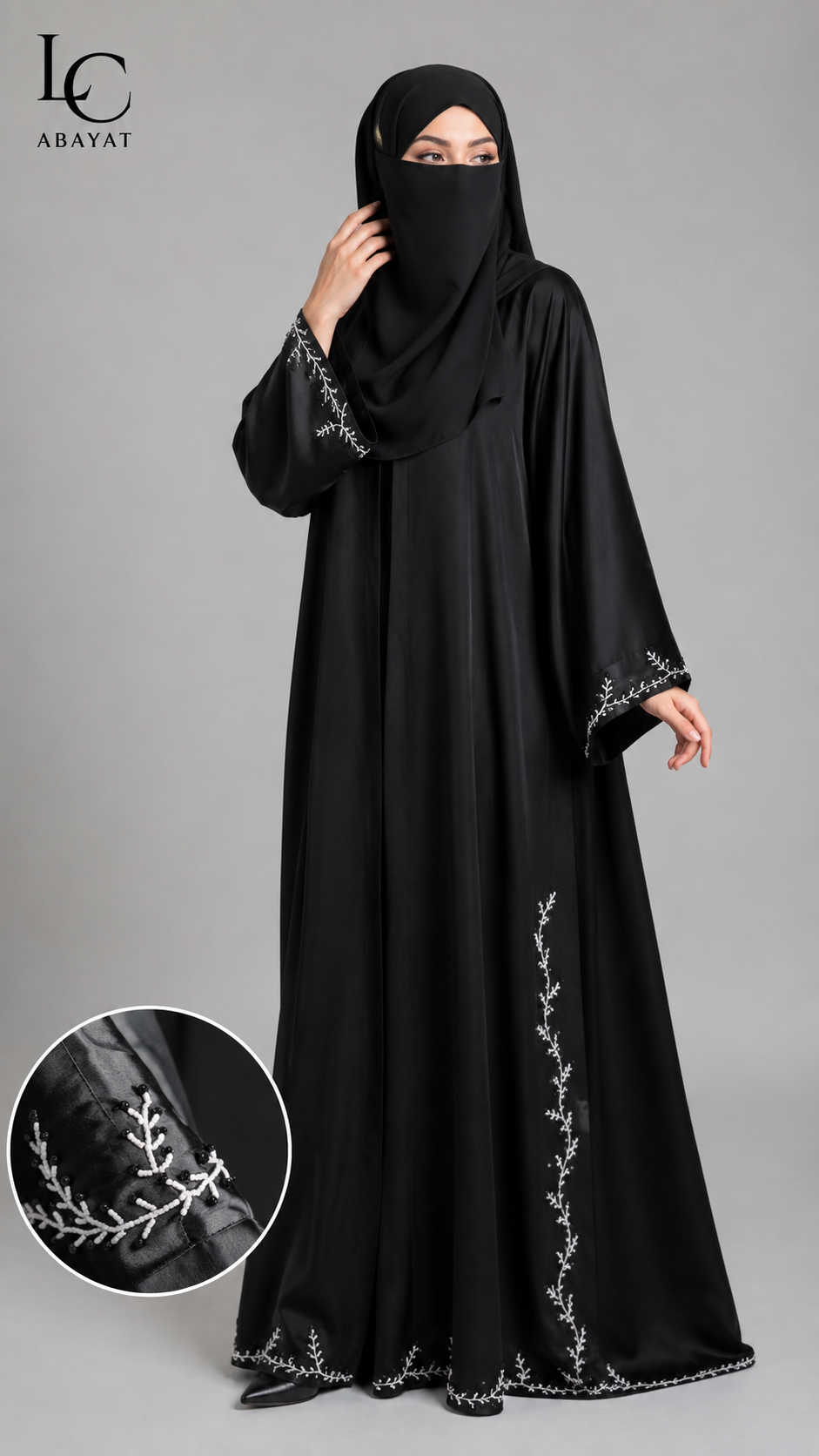 A woman wearing a black abaya with silver embroidered details and a matching black hijab, shown against a gray studio background. A circular inset at the bottom left highlights the silver embroidery on the sleeve.