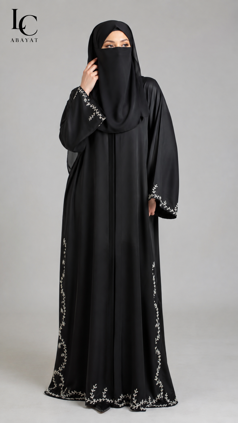 A woman wearing a black abaya with silver floral embroidery on the sleeves and hem, paired with a matching black hijab. She is standing against a neutral gray studio background.