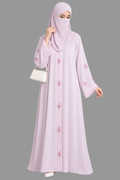 A woman modeling a light lavender cutdana embellishment hanwork modest abaya, including a long abaya with pink floral embroidery, a matching headscarf, and a small white handbag, on a white background with the Abayat Limited Collection brand logo in the top left corner.