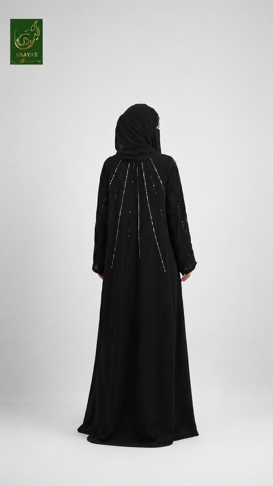 A woman wearing a long black abaya and matching black hijab, shown from the back against a light gray studio background. The abaya features delicate silver beaded vertical lines down the center back.