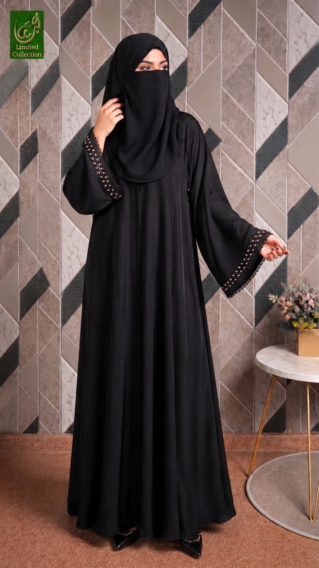 A woman wearing a black abaya with decorative gold chain trim on the sleeves and a matching black hijab, standing in front of a geometric tiled wall.